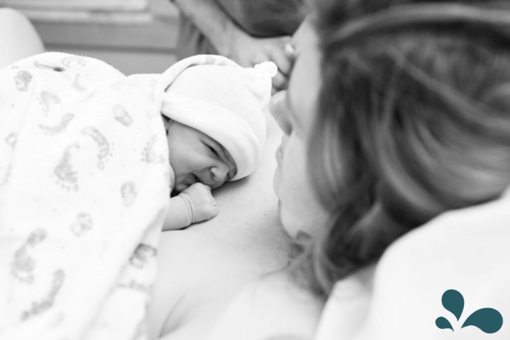 Black and white photo of a newborn in a hat resting on its mother's chest for skin-to-skin contact immediately after birth, both appear peaceful.