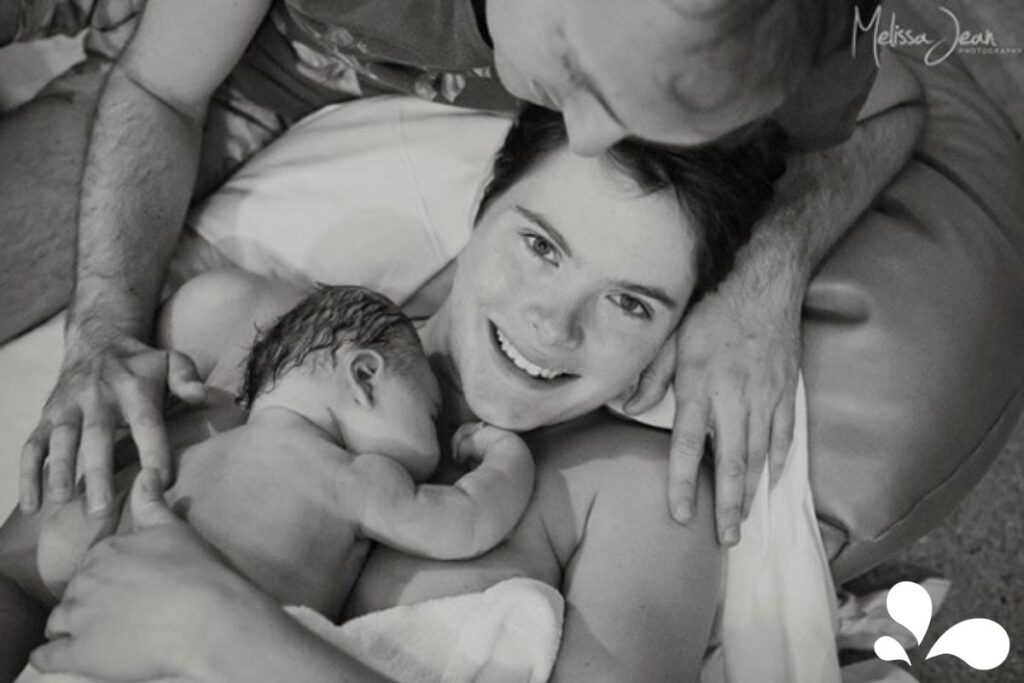  black and white photo of a joyful mother in a hospital bed, smiling up at her partner who is leaning over her, as they both lovingly embrace their newborn lying on the mother's chest, experiencing the warmth and comfort of skin-to-skin contact.