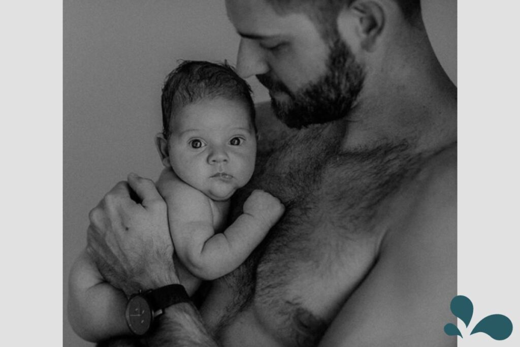 A black and white photo capturing a tender moment where a newborn baby, wide-eyed and alert, is held securely in the arms of their bearded father, both skin-to-skin, enhancing bonding.