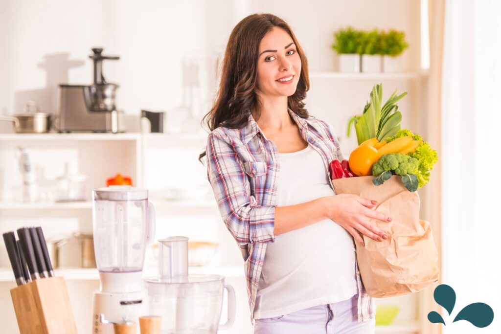 A smiling expectant mother holding a paper bag full of fresh produce including vegetables and fruits, standing in a modern kitchen