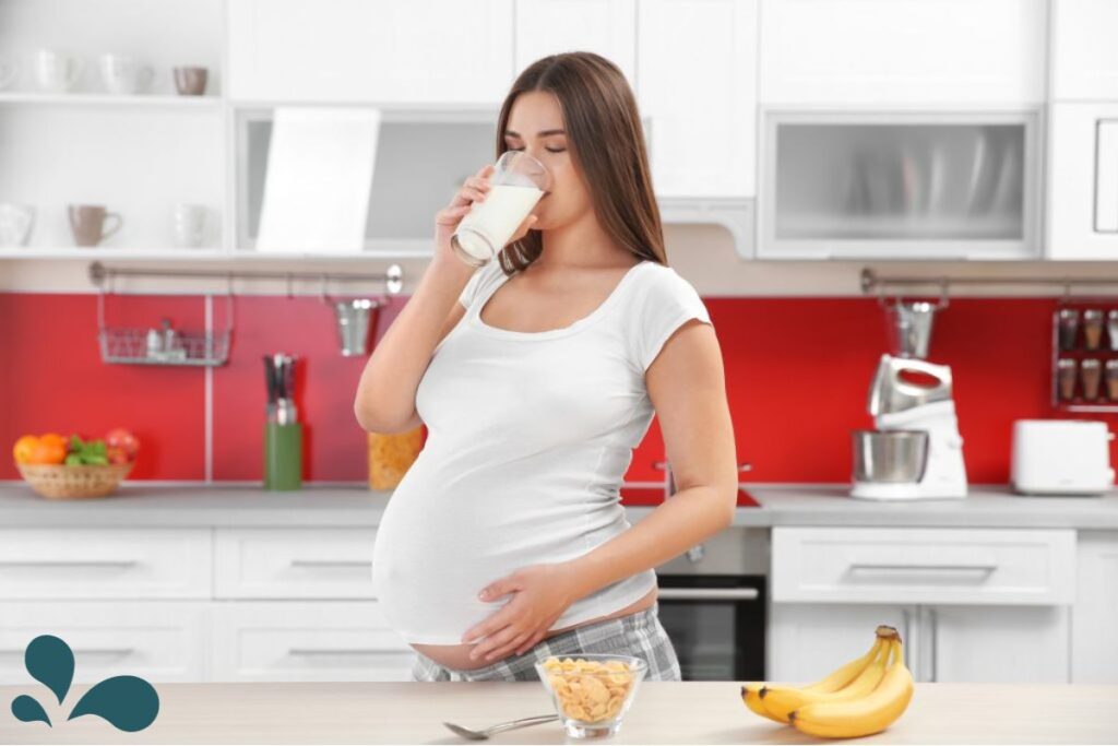 Pregnant woman in a white top enjoying a nutritious breakfast of cereal and milk with bananas on the kitchen counter, promoting a healthy pregnancy diet.