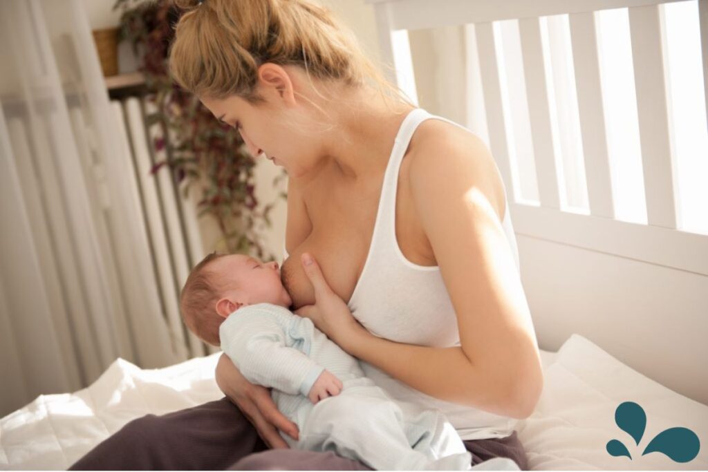 Mom breastfeeding her baby in a white tank top while sitting by the window on her bed.