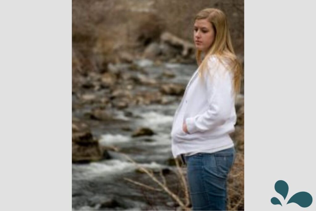 A thoughtful woman stands by a riverbank in a serene natural setting, lost in contemplation with water flowing in the background.