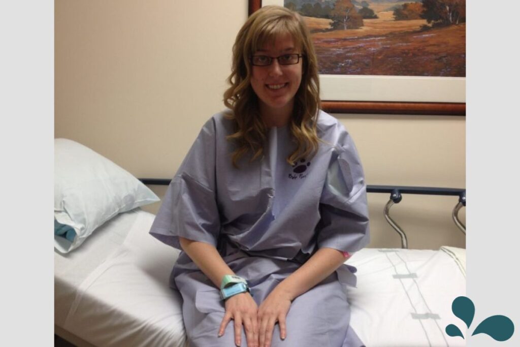 A woman sits on a hospital bed wearing a patient gown and a wristband, smiling before a medical procedure, with a calm demeanor.