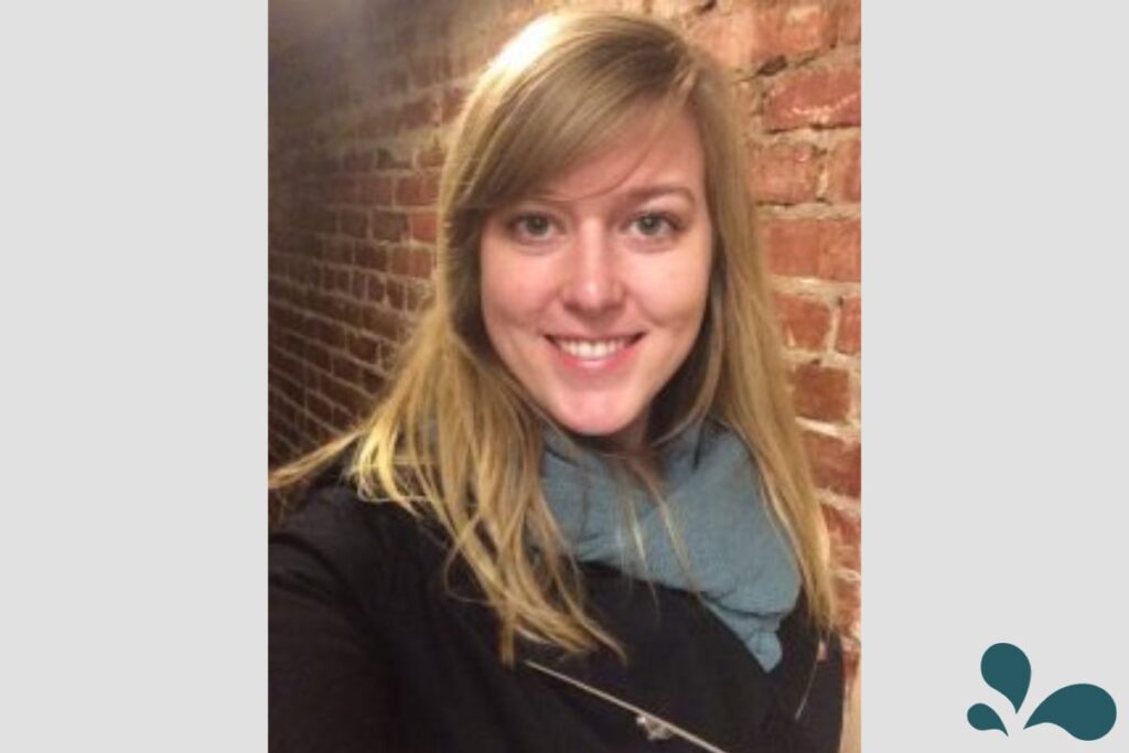 A woman in a black jacket and teal scarf smiles for a selfie, with the warm ambiance of indoor lighting and a brick wall background.