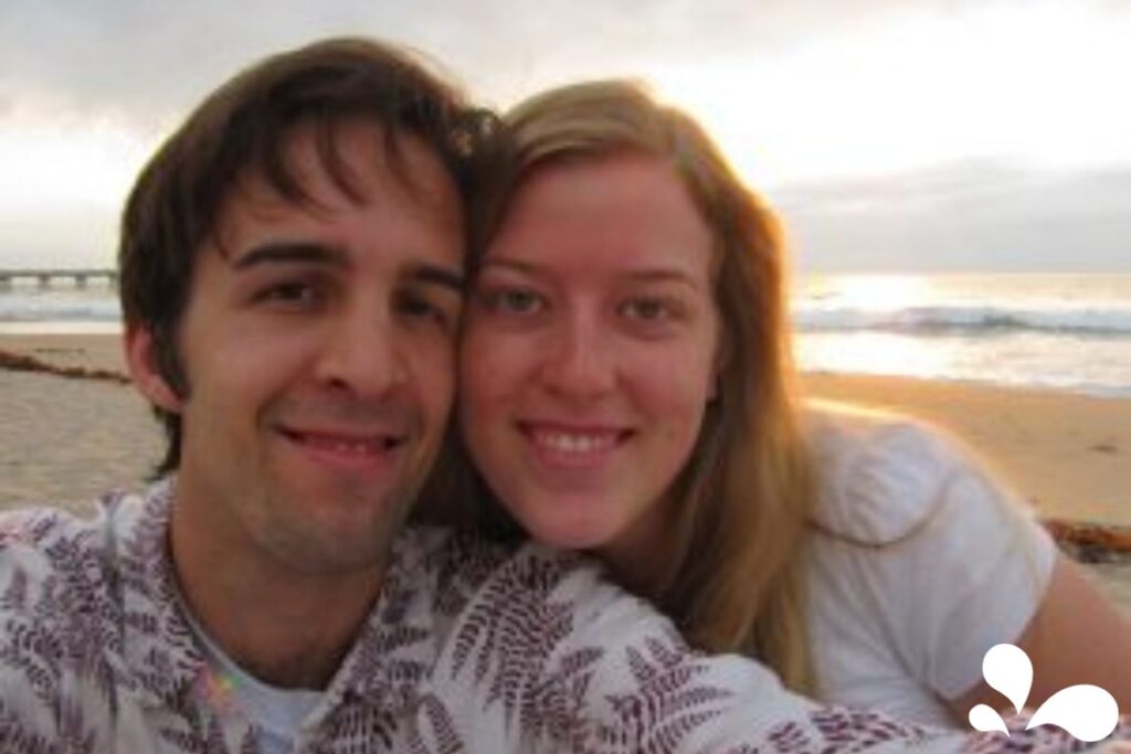 A couple takes a selfie with a glowing sunset over the ocean behind them, sharing a smile and a close embrace on the beach.