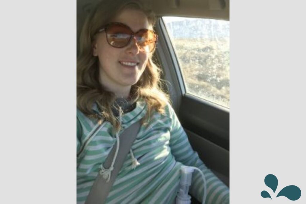 A woman in a striped hoodie and sunglasses, using a breastpump, smiles while driving, captured in a car interior during the day.