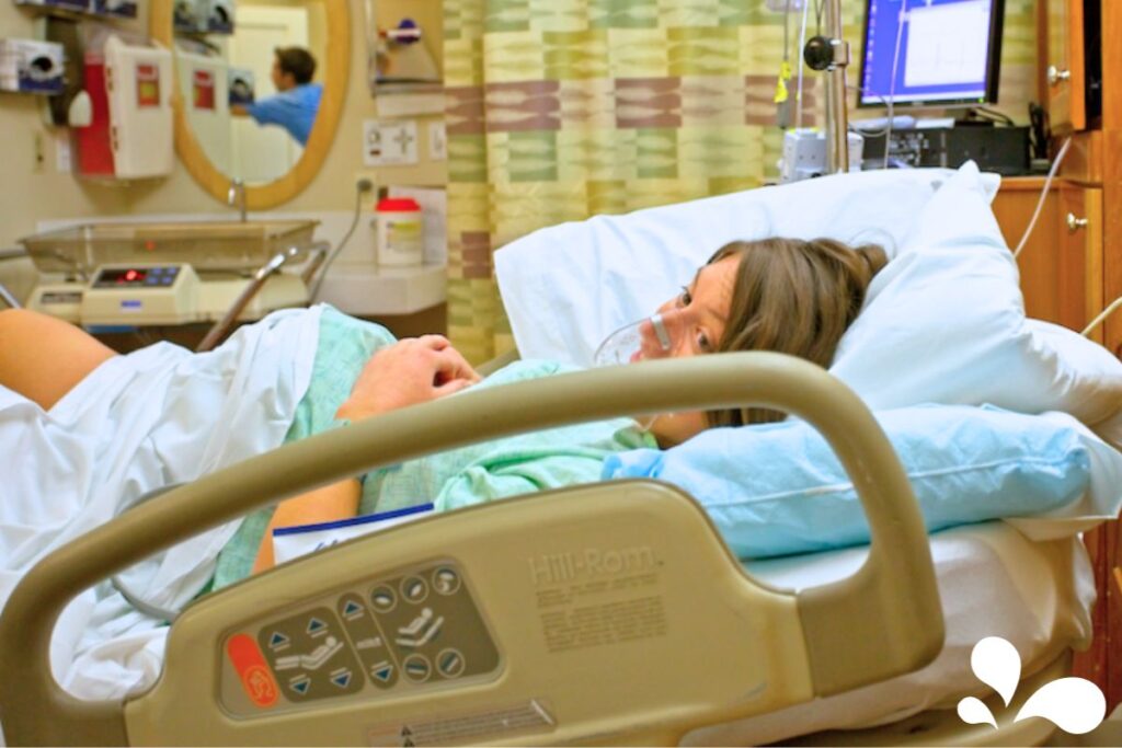 A woman in labor breathing through contractions with an oxygen mask in a hospital delivery room, surrounded by medical equipment.
