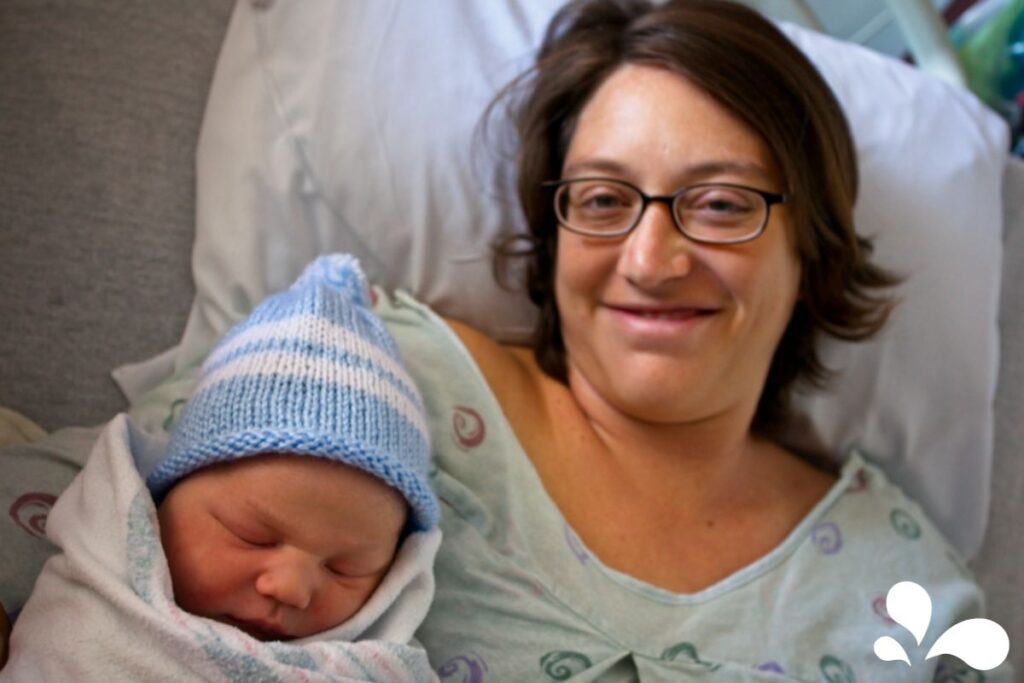 A happy new mother in a hospital gown, wearing glasses, holds her newborn baby sleeping in a blue striped hat