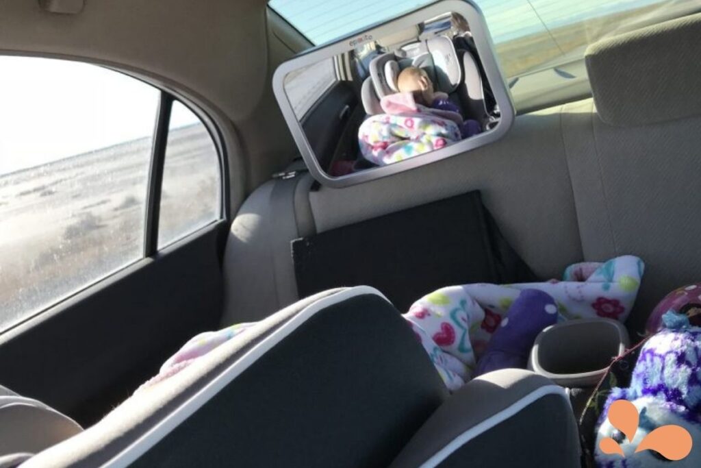 A rear car seat view showing a baby in a colorful car seat through a reflection in the mirror, surrounded by plush toys and blankets.