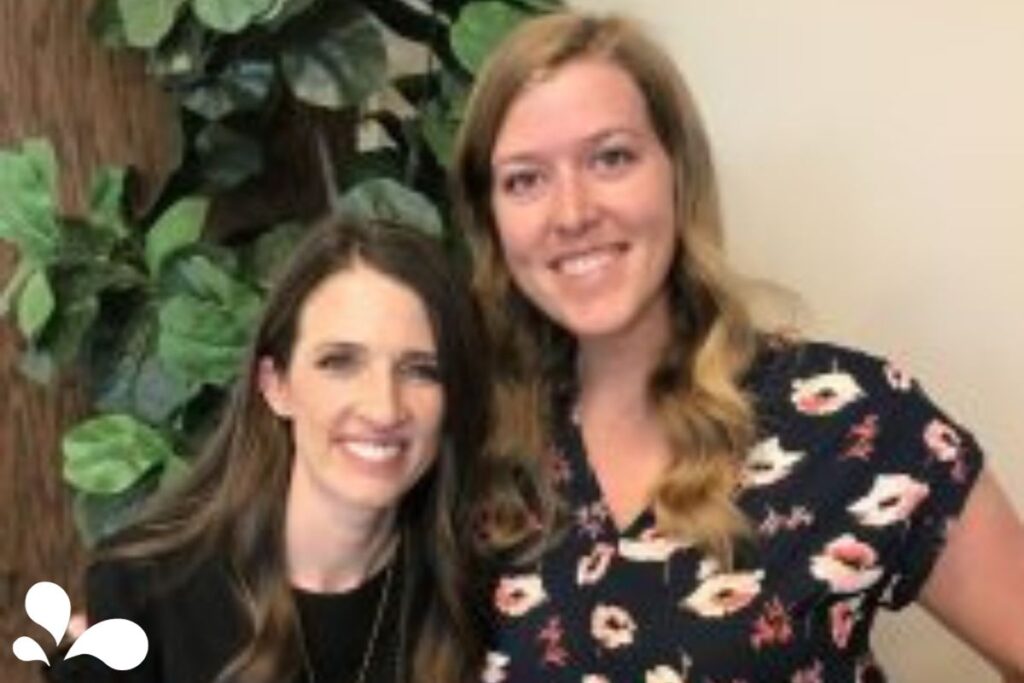 Photograph of two women standing side by side, smiling at the camera. The woman on the left has long straight dark hair and wears a black top, and the woman on the right has long wavy brown hair and a floral dress. They are in front of a backdrop of indoor greenery.