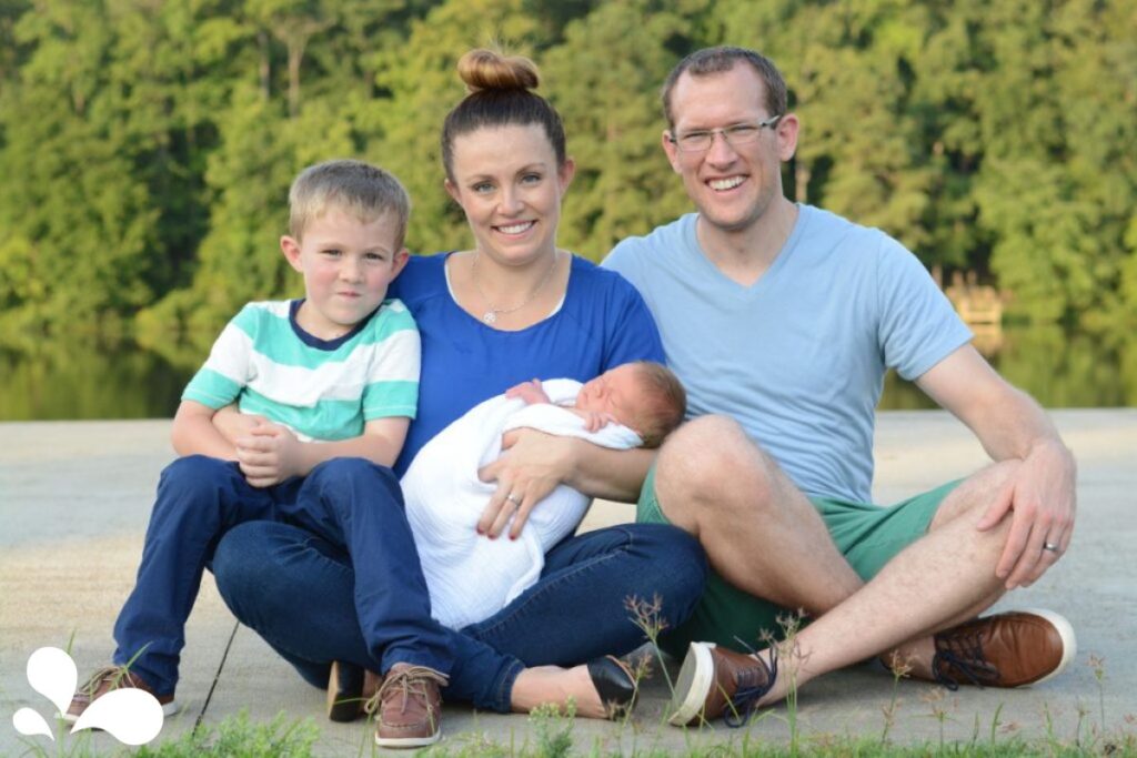 The Dewey family is seated outdoors, enjoying a serene moment by a lake. A father in a blue tee and green shorts, a mother in a navy dress, cradling a sleeping newborn wrapped in a white blanket, and their older son in a green and white striped shirt and navy pants, all smile warmly at the camera. The backdrop of lush green trees and calm water reflects the tranquility and happiness of their family life.