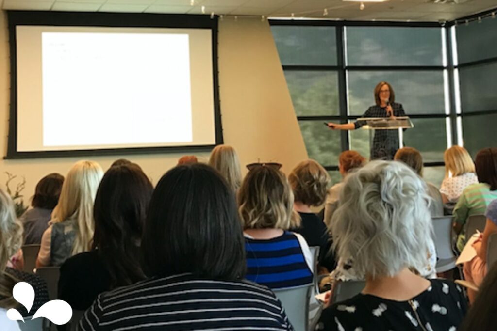 A speaker at a conference, standing at a podium and gesturing towards a presentation on a large screen, in front of an audience of attentive listeners.