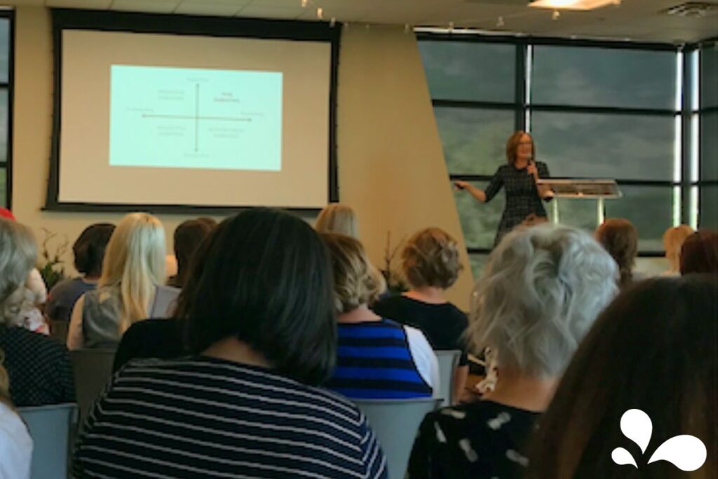 A presenter at a lectern pointing to a parenting style graph projected on a screen in a conference room filled with attentive audience members. The graph shows different parenting styles categorized along axes of 'Supportive' and 'Demanding.