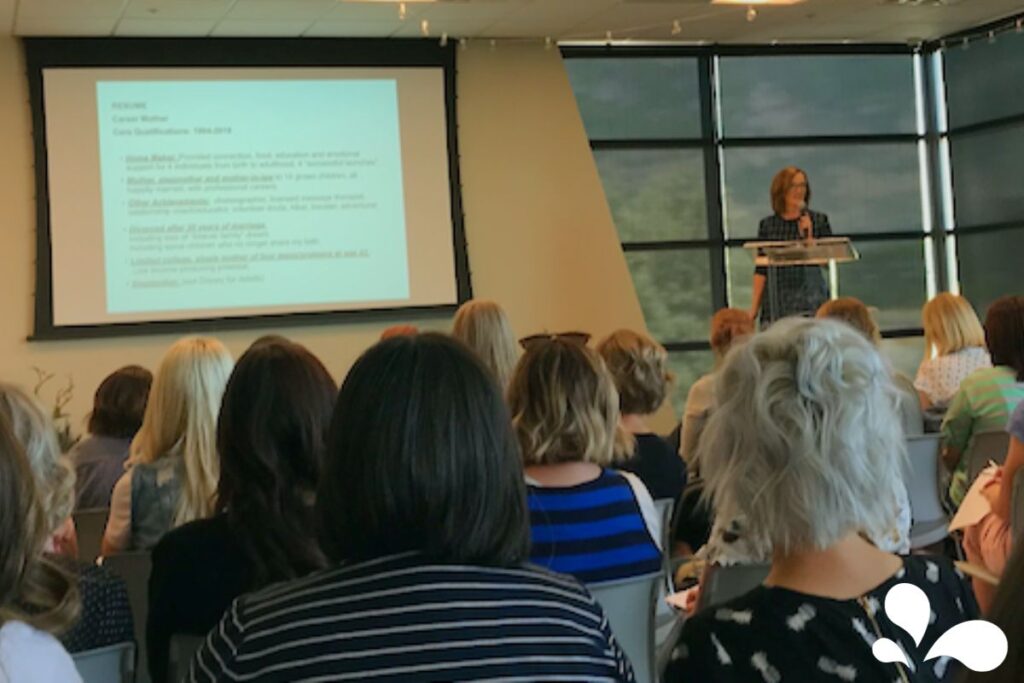 A speaker at a podium addressing an audience during a conference. The speaker appears to be presenting a resume on a projection screen in a room with large windows, while attendees listen attentively.