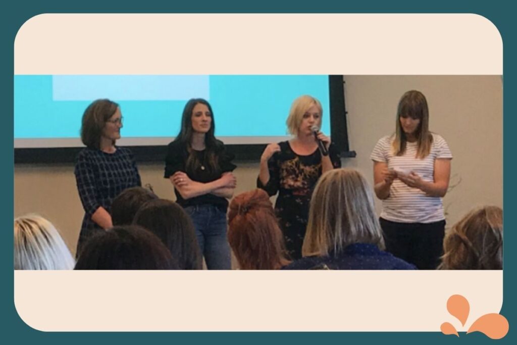 Image of Georgia Anderson, Ralphie Jacobs, Jody Moore, and Lisa Andersen standing together at a conference, with Georgia speaking to an audience and the others listening and observing.