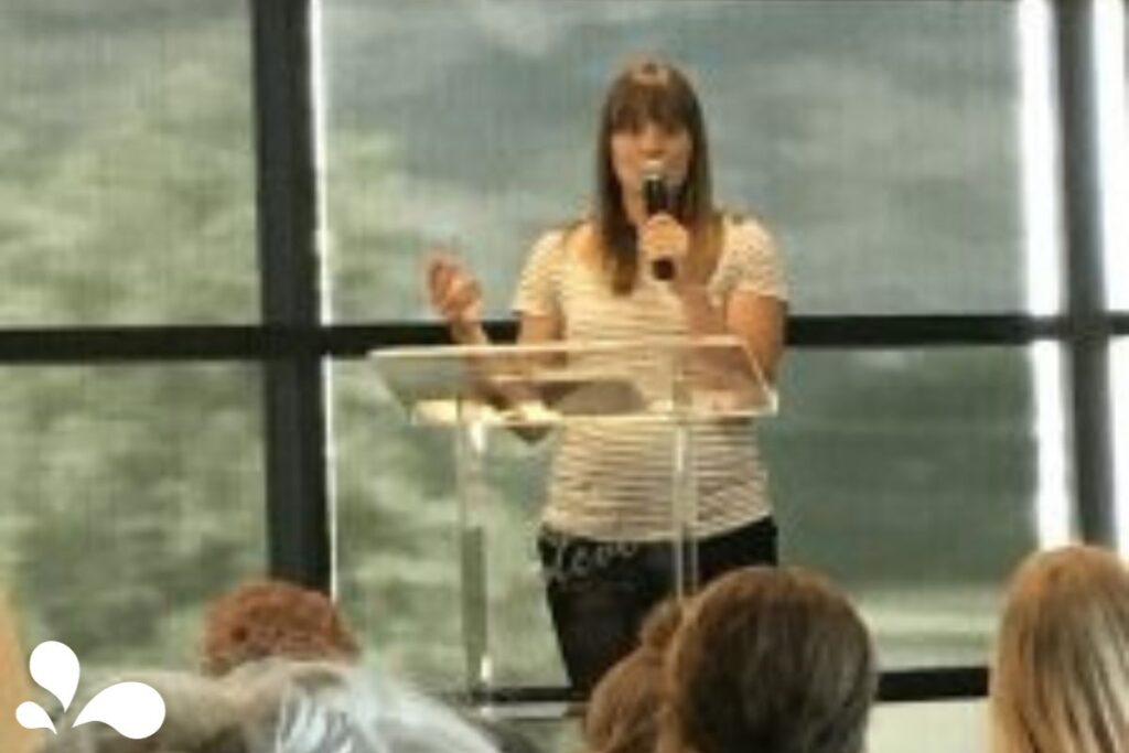 A woman with shoulder-length hair speaking into a microphone at a podium, addressing an audience in a room with large windows and natural light.