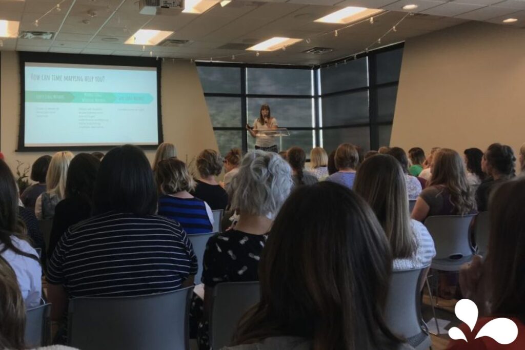 A speaker at a podium in a conference room full of attendees, with a presentation slide titled 'How Can Time Mapping Help You?' visible in the background.