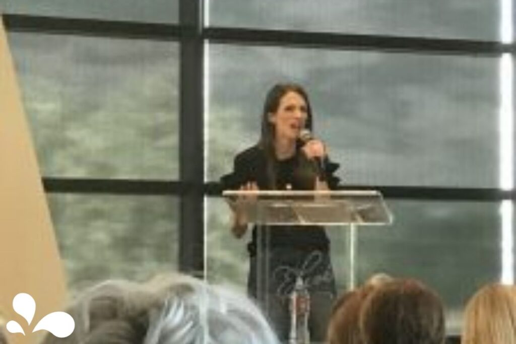 Image of a woman speaking at a podium in a conference room with large windows in the background. The audience is in the foreground, out of focus. The speaker has long dark hair, wearing a black top, and is gesturing with her right hand.