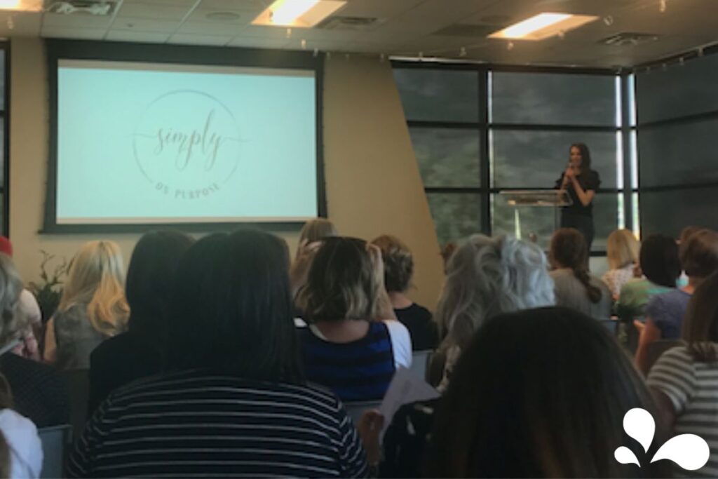 Image of Ralphie Jacobs giving a presentation in a conference room with a large audience in attendance. A projection screen behind her displays the text 'simply on purpose'. She is standing at a clear podium, addressing the audience with a microphone, with large windows allowing natural light into the room.