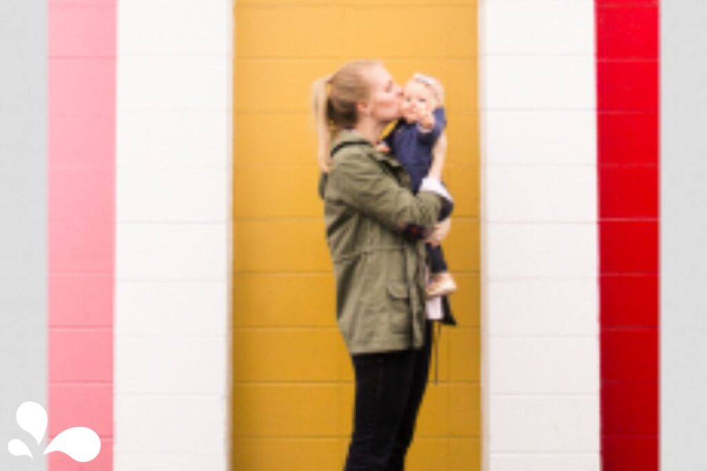A candid snapshot of Adrianne lovingly kissing her baby against a vibrant, multicolored striped wall. Adrianne, dressed in a casual olive green jacket and her hair in a ponytail, holds her baby who wears a cute navy outfit and seems to be enjoying the affectionate gesture. The bold yellow, white, and red stripes on the wall add a playful and artistic backdrop to this sweet mother-child interaction.