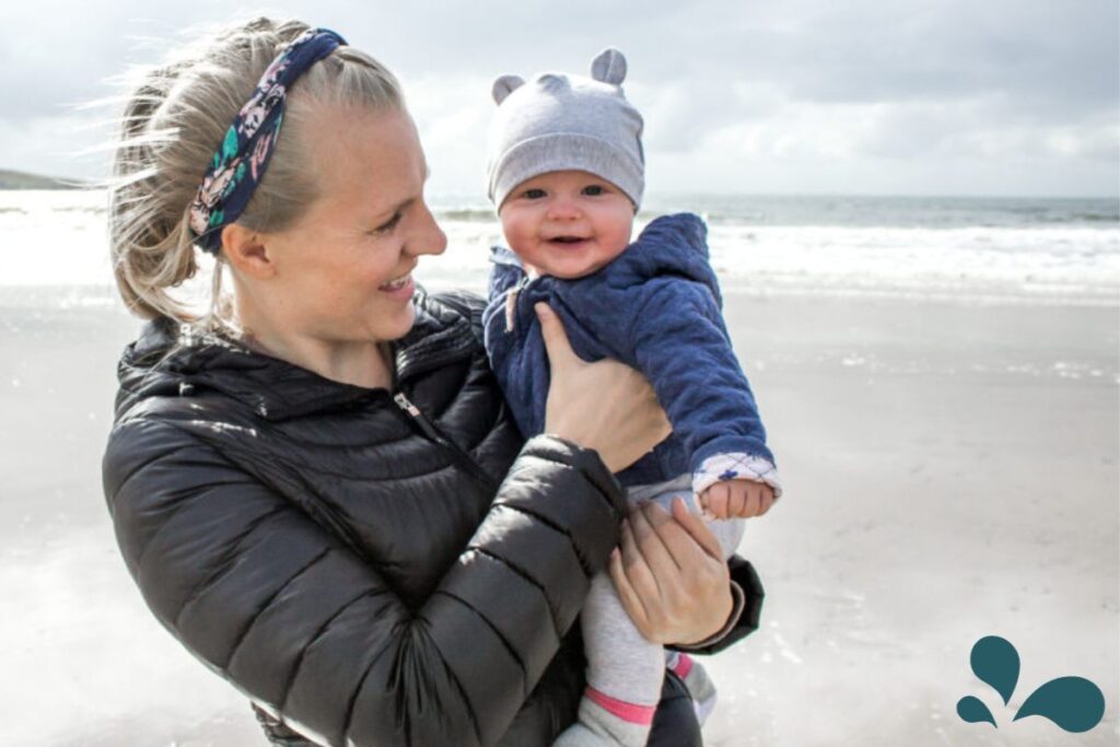 A joyful mother in a black jacket and blue headband holds her smiling baby, who is bundled up in a blue jacket and cute grey bear-eared hat, at the beach. The background shows a serene beachscape with waves gently lapping at the shore, under a cloudy sky. This image captures the happiness and loving bond shared during a day out at the beach.