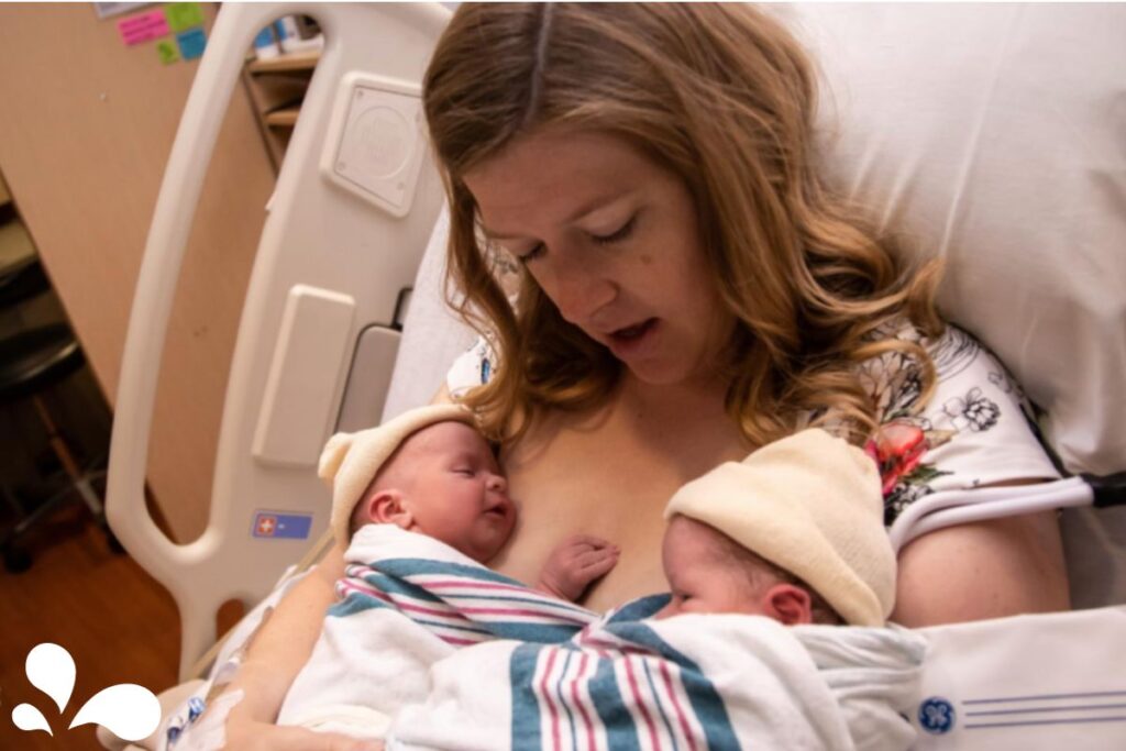 A mother sitting up in a hospital bed looking down fondly at her two newborns cradled on her chest for skin-to-skin time.