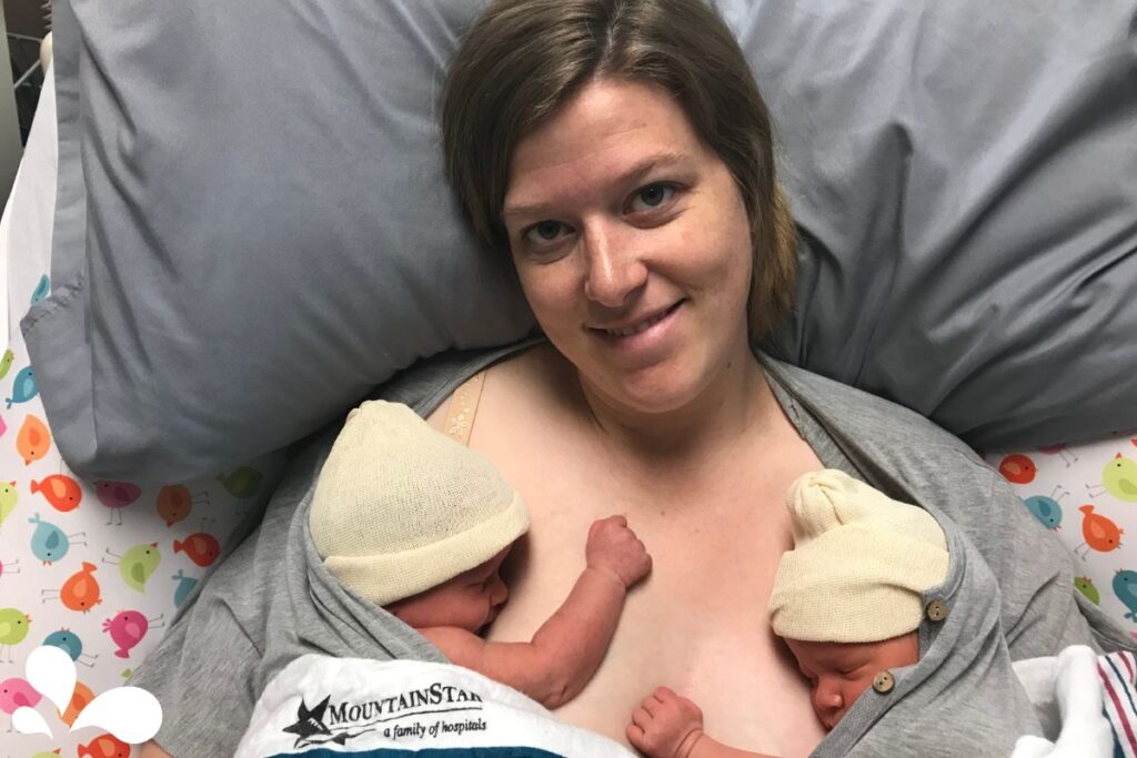 A new mother smiling at the camera while practicing skin-to-skin contact with her twin babies in a hospital setting, each child snug in a soft hat.
