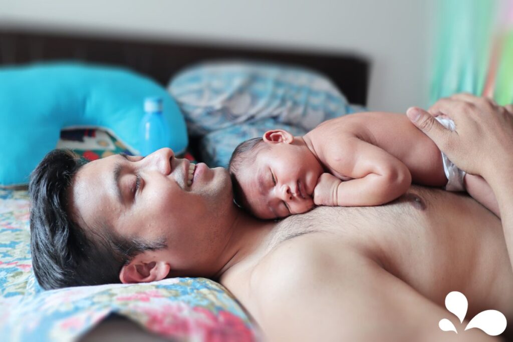 A father lying back with eyes closed, enjoying the tranquility of skin-to-skin bonding with his sleeping newborn resting on his chest.
