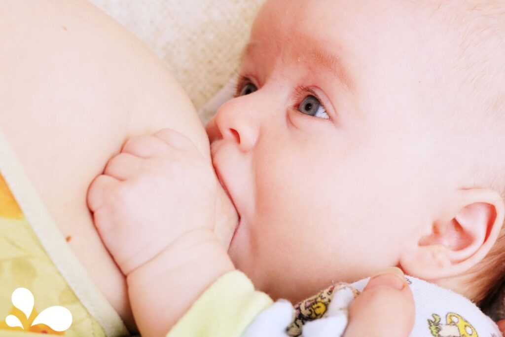 A close-up image of a breastfeeding baby with wide, curious eyes looking upwards. The baby's hand is gently resting on the mother, creating a powerful image of the connection and trust involved in breastfeeding.