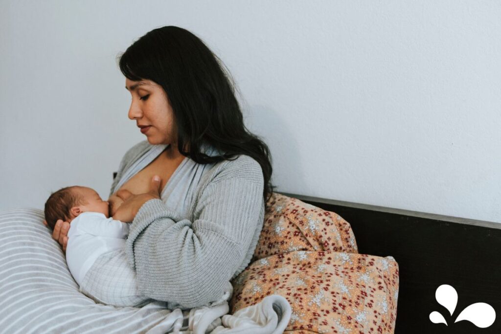A mother, seated comfortably and breastfeeding her baby, appears calm and reflective. The coziness of the room and the mother's gentle hold around her baby illustrate the quiet moments of motherhood and the intimate experience of breastfeeding.