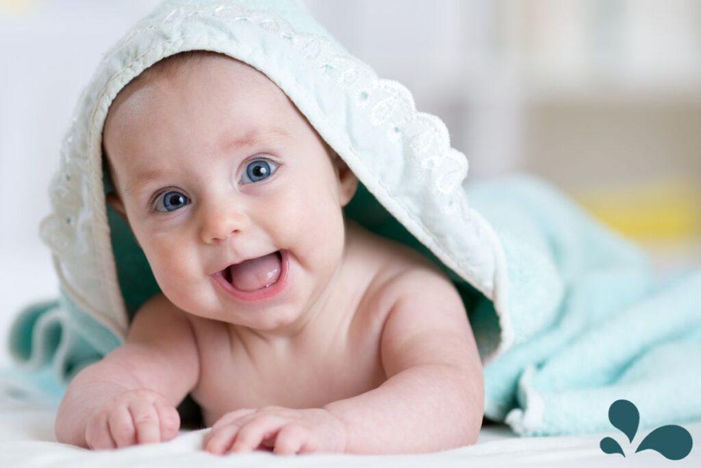 A joyful baby peeking out of a hooded towel with a lace trim, showing bright blue eyes and a wide smile.