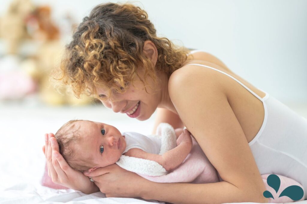 A mother with curly hair cradles her newborn in her arms while lying on a bed, both sharing a tender gaze.
