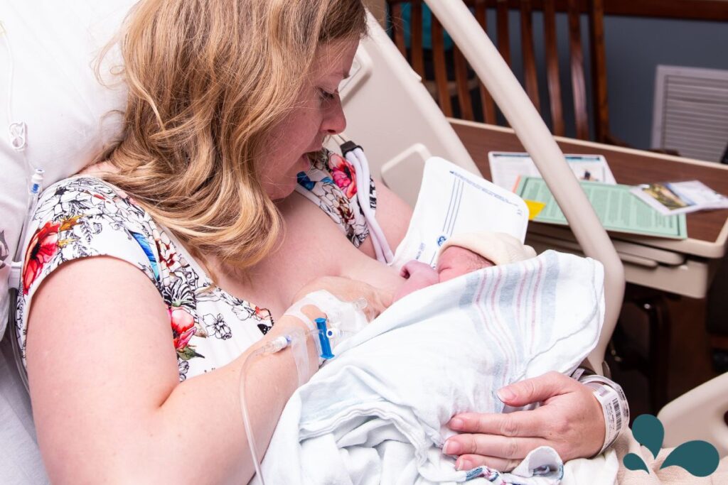 A mother in a hospital bed lovingly breastfeeding her newborn, with educational cards in the background.