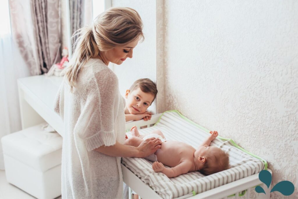 A mother changing her baby on a changing pad, with an older child looking on with affection, in a warmly lit, comfortable nursery.