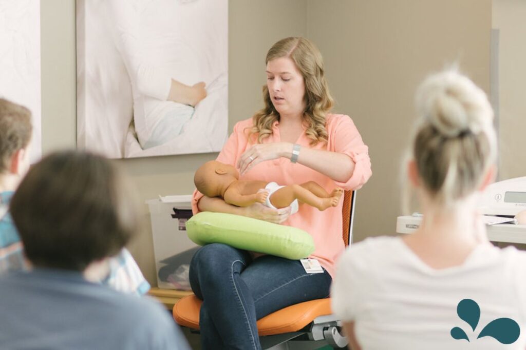 An IBCLC is demonstrating how to properly position a baby to prepare to breastfeed. The class is filled with expecting parents who are eagerly learning breastfeeding knowledge in a bright, cozy classroom setting.