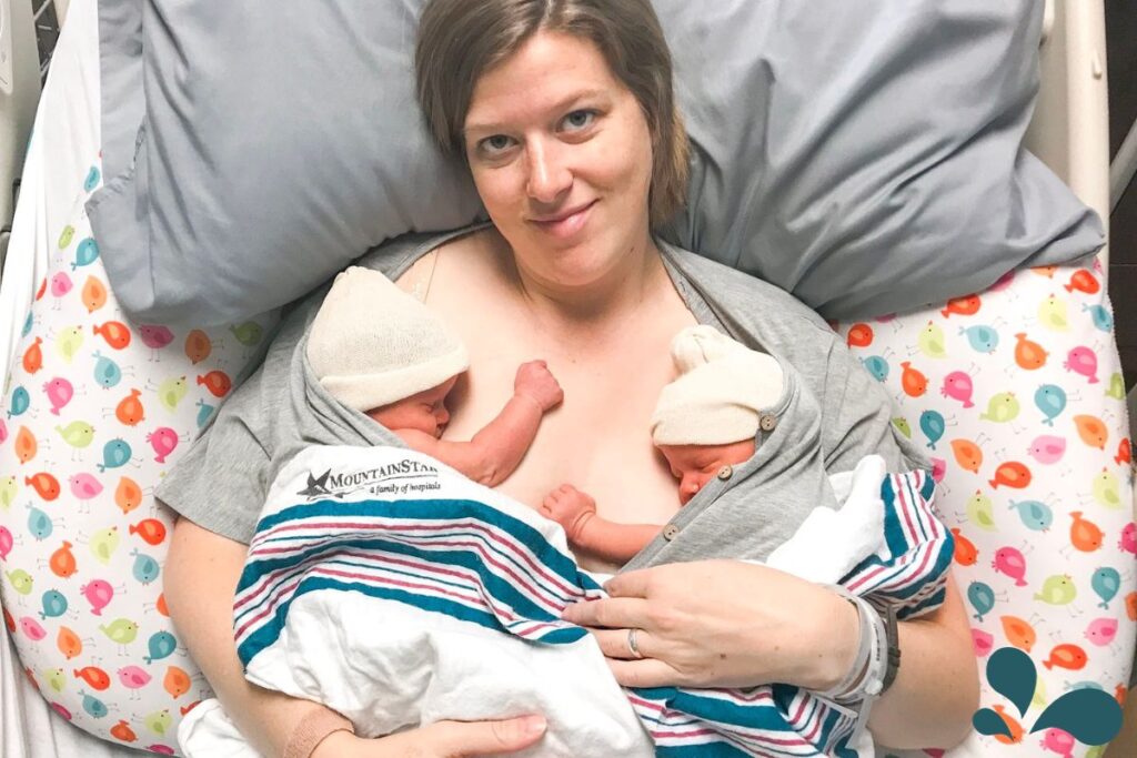 Content mom holding newborn twins in her hospital bed. Twins and mom are skin-to-skin while covered with a white and striped blanket over the top of them.