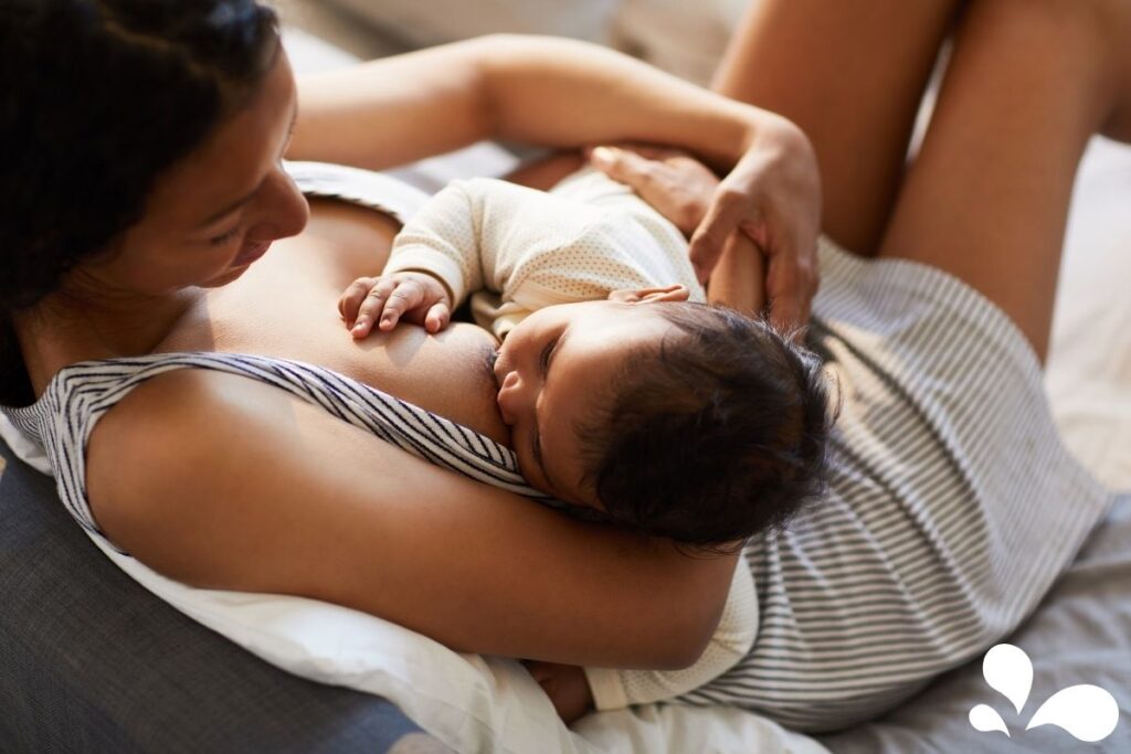 A serene image of a mom lying back and breastfeeding her baby while resting on the couch. Peaceful bonding of mom and baby with the soft light shining behind them.