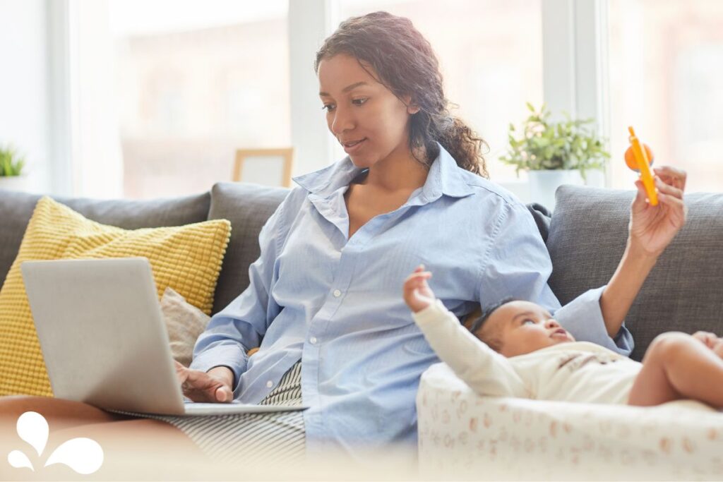 An image of a mother working from home on her laptop with her baby playing beside her, depicting the new normal for working mothers.