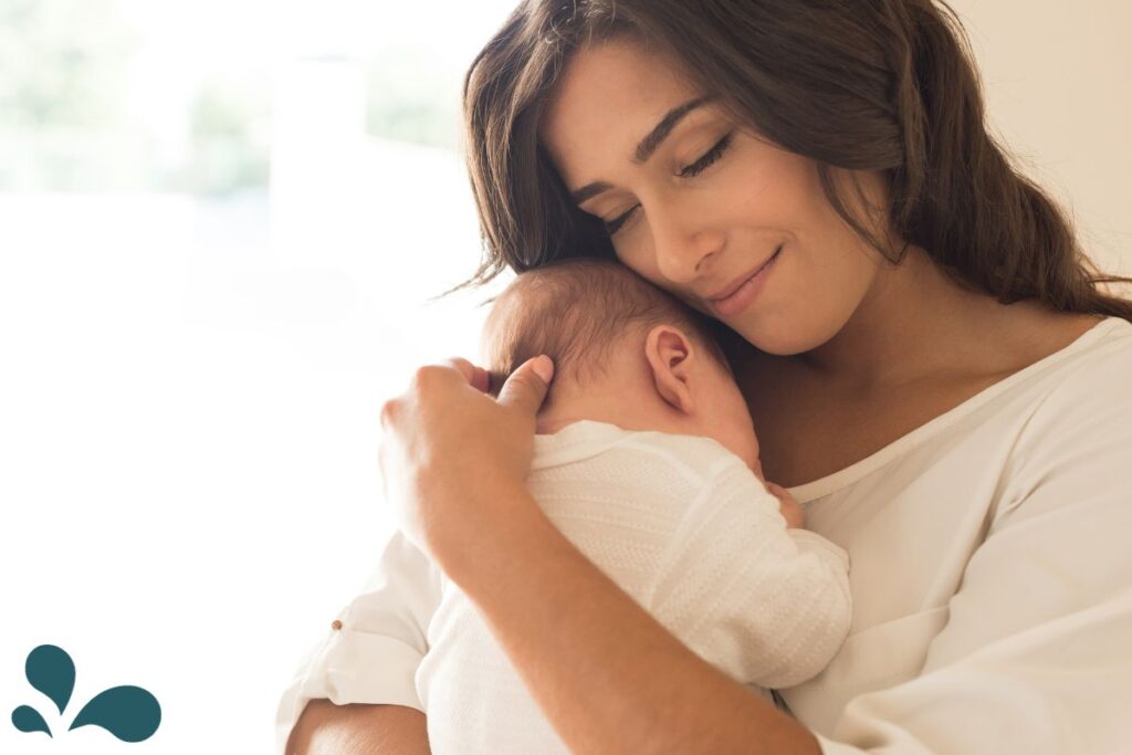A serene mother with a gentle smile cradles her sleeping newborn close to her chest. The baby is wrapped in a delicate white blanket, suggesting warmth and comfort. This intimate moment is captured in a softly lit room, conveying a sense of peace and the deep emotional connection between parent and child.
