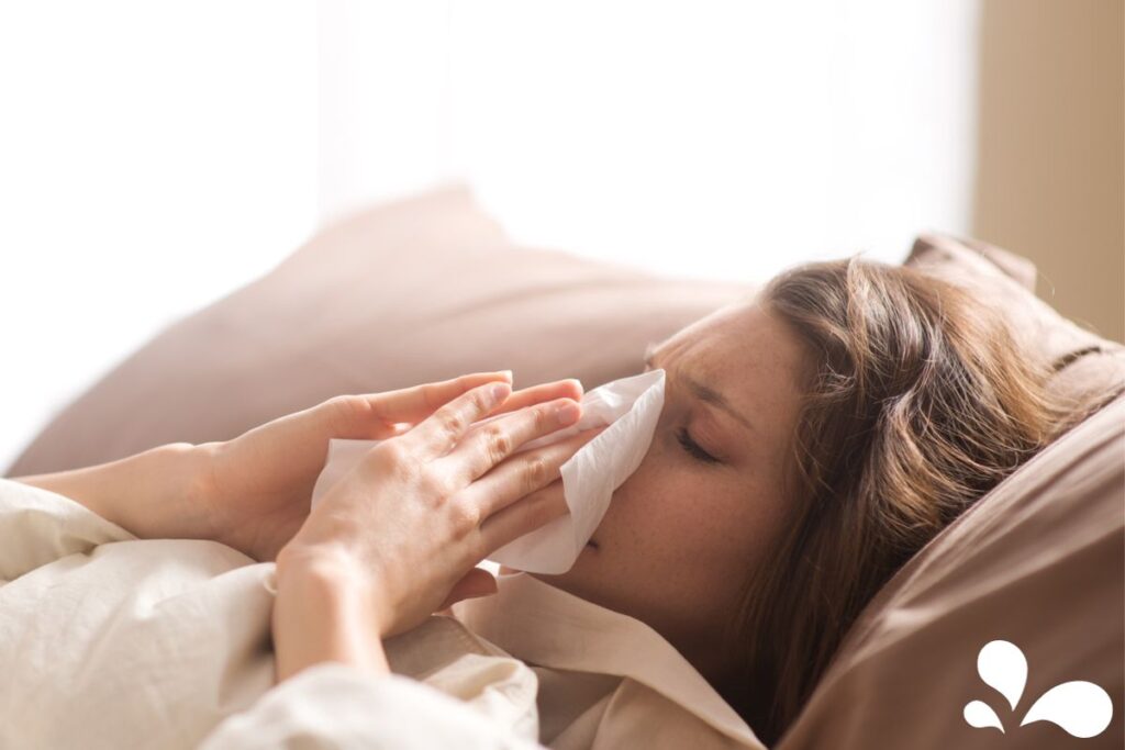 A woman is lying in bed, her face partly obscured as she blows her nose with a tissue, a common self-care practice during a cold or flu. The lighting is soft and warm, suggesting a quiet moment of rest and recovery in the comfort of her bedroom.