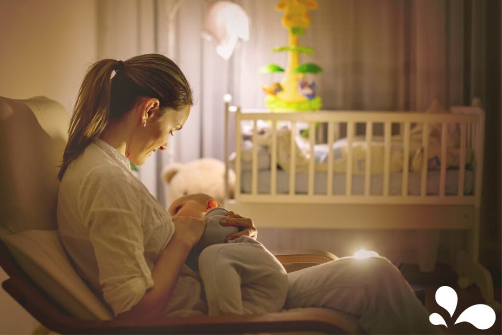 A mother sits in a rocking chair, tenderly breastfeeding her baby in the soft glow of evening light. They are in a cozy nursery, with the baby’s crib and toys in the background, creating a peaceful and intimate atmosphere for this nighttime feeding moment.