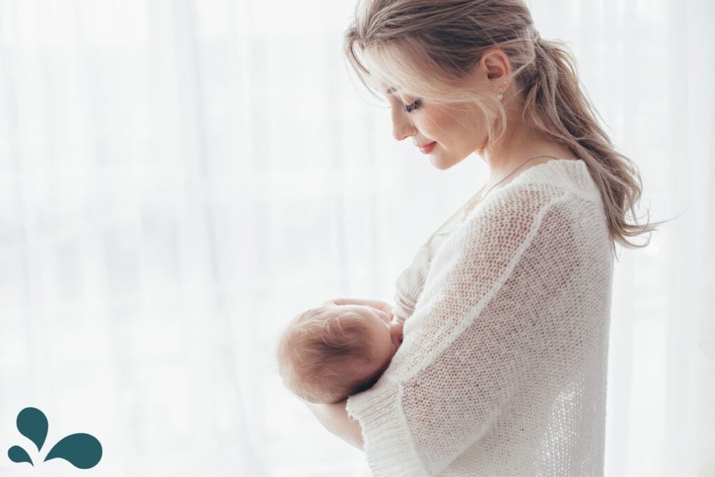 A serene mother in a white knitted sweater lovingly cradles her breastfeeding infant, bathed in the soft light of a bright window, creating a peaceful and intimate atmosphere.