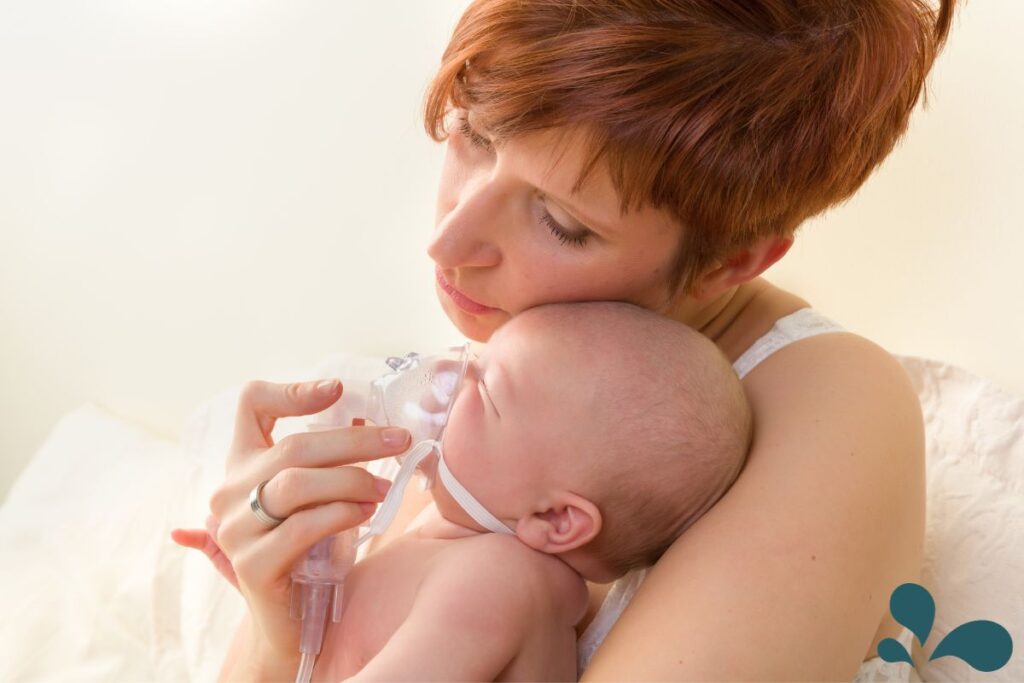 A mother gently holds her baby, providing a breathing treatment with a small, handheld nebulizer. The baby is calm, nestled against her, suggesting trust and comfort even during medical care. The mother's eyes are closed in a moment of tenderness, her expression soft and loving, as sunlight gently illuminates the quiet scene.