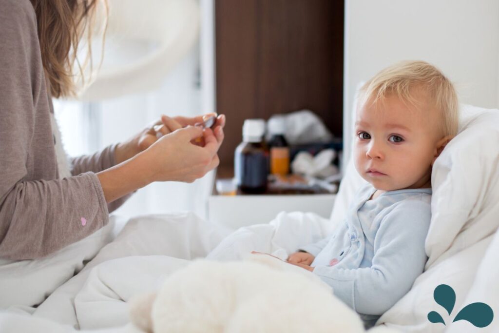 A concerned mother prepares medication for her blonde toddler who is propped up in bed, looking unwell and weary. The background is a soft-focused home setting with medicine bottles, suggesting a caring moment during a child's illness. The image captures a common parenting experience of tending to a sick child with gentle care.
