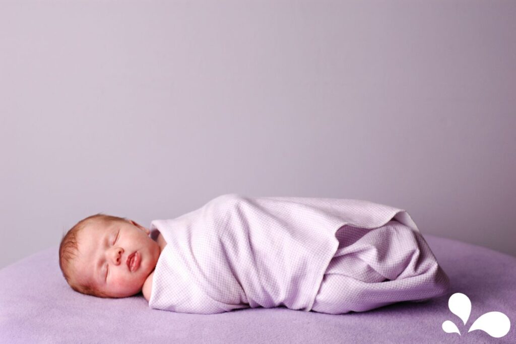 A newborn baby peacefully asleep, swaddled in a light purple blanket on a soft purple surface. The baby's gentle expression and relaxed posture capture the essence of newborn tranquility and contentment.