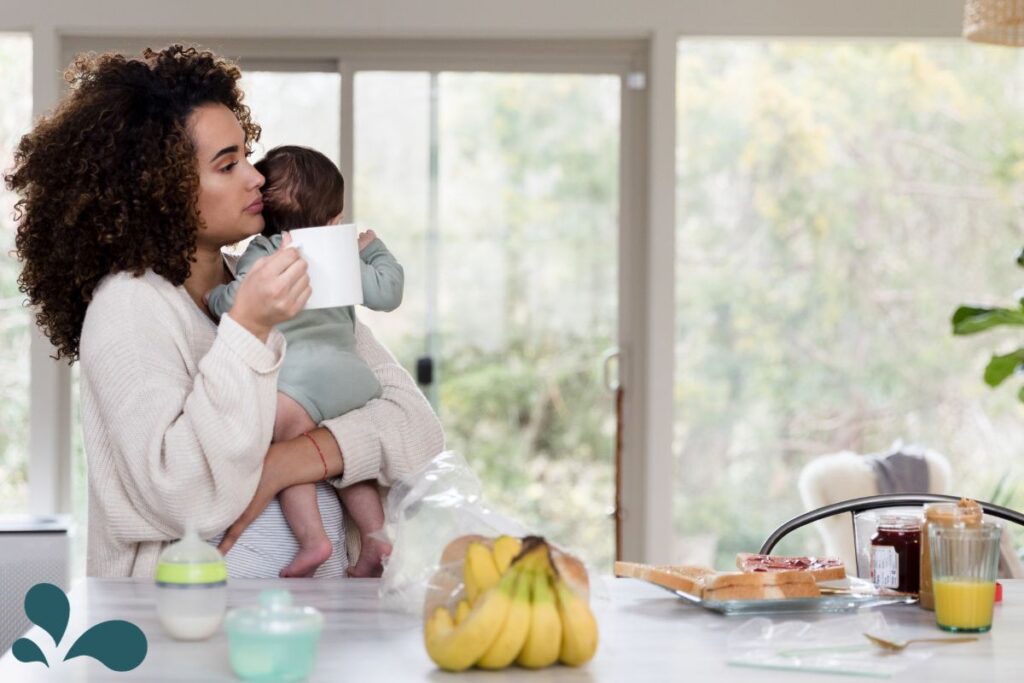 A mother lovingly holds her baby close while enjoying a cup of coffee in a sunlit, cozy kitchen. The breakfast table is casually set with a loaf of bread, a jar of jam, and a glass of juice, evoking a sense of warmth and domestic tranquility. Large windows reveal a serene, natural outdoor backdrop, adding to the peacefulness of the moment.