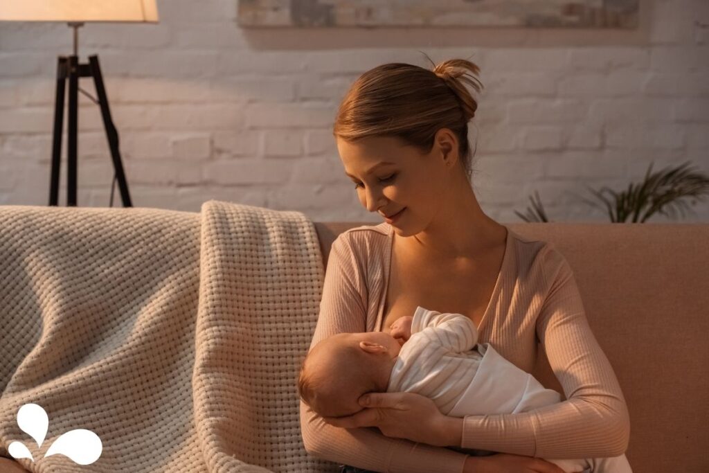 A serene moment as a mother sits on a beige couch, gently cradling and breastfeeding her newborn. She gazes affectionately at her baby, enveloped in a soft white blanket, under the warm glow of a lamp in a cozy room with a white brick wall in the background, symbolizing the intimate and nurturing bond between parent and child.