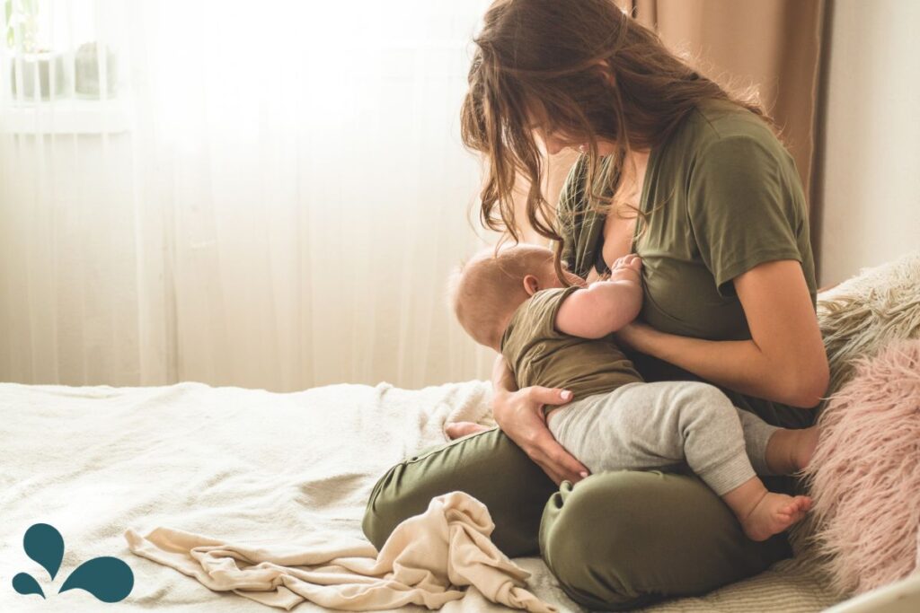 A tender moment of motherhood is captured as a woman in a matching olive green top and pants breastfeeds her baby, who is also dressed in green. They are sitting on a bed with a white blanket, near a fluffy pink pillow, with soft natural light streaming in through the window, creating a serene and intimate atmosphere.