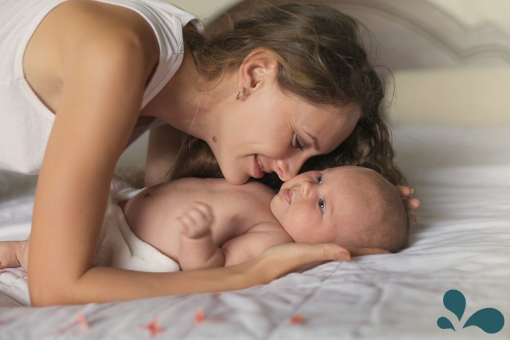 A tender scene of a mother in a white tank top as she lovingly gazes down at her newborn, lying on a bed with a white and star-patterned blanket. The baby looks back with trust and affection, illustrating the unique and loving bond between a mother and her child in a serene and comfortable bedroom setting.